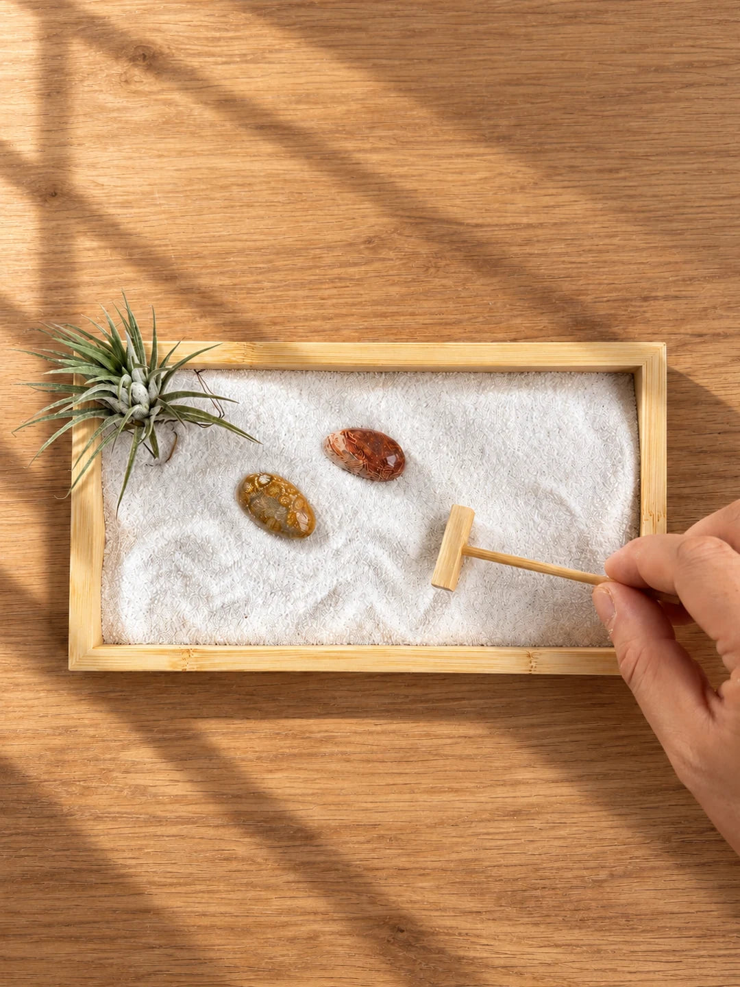 A hand holding a wooden sand tool above a bamboo Zen garden with white sand, two patterned stones and an air plant, lit by soft diagonal window sunlight on a wooden table; portrait banner.