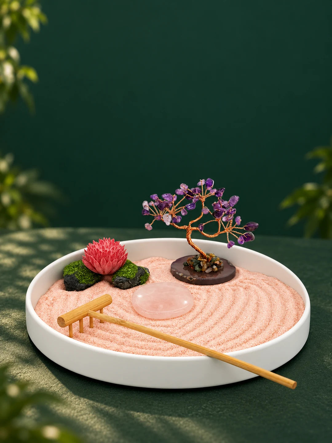 A white round Zen garden tray with peach-pink sand, a purple crystal tree, pink lotus ornament, pale pink polished stone, and bamboo rake on a forest-green surface; portrait banner.