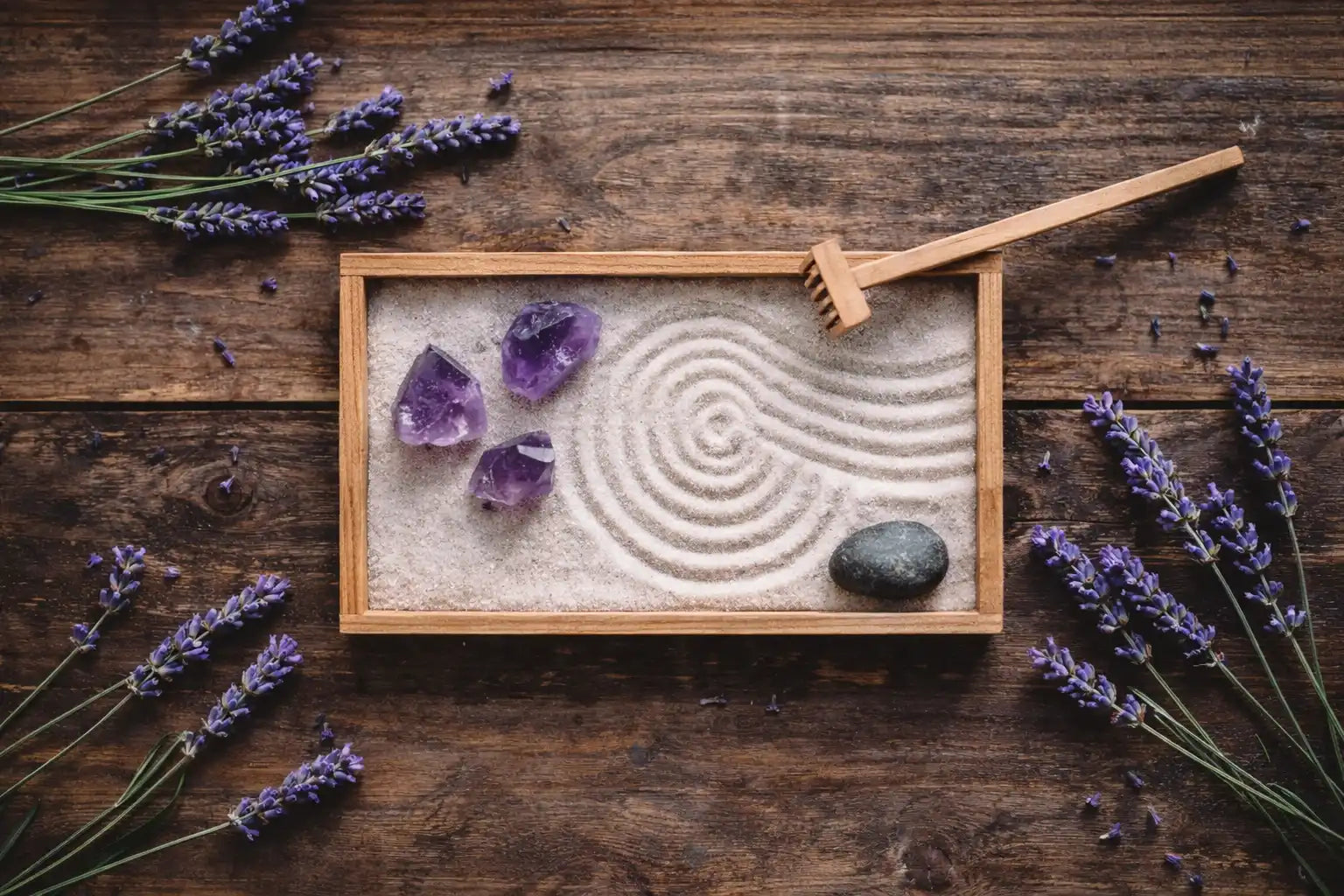A top-down photograph of a small zen garden on a wooden table, featuring white sand with raked patterns, raw amethyst crystals, lavender sprigs, a smooth stone, and a wooden rake under soft natural light.