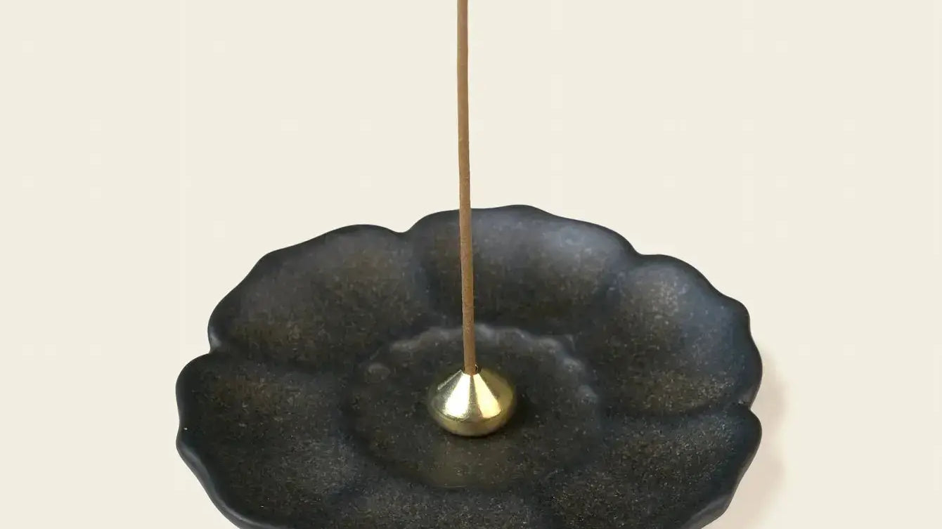 Dark ceramic lotus-shaped incense tray with a small brass holder and an unlit incense stick.