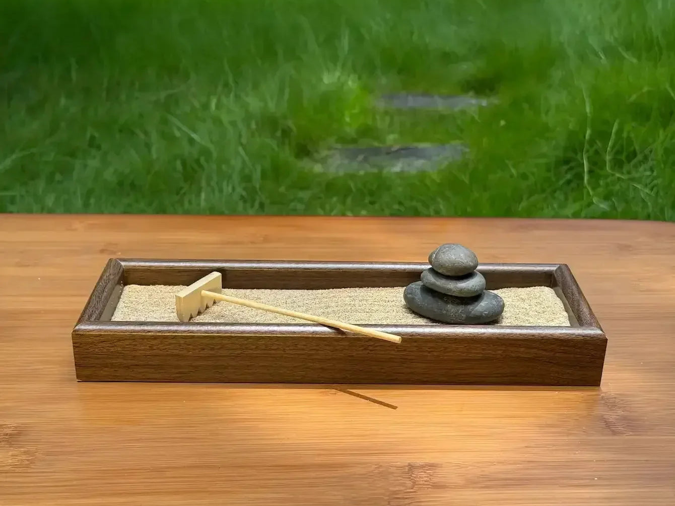 Wooden tabletop garden with three stacked river stones, sand, and a bamboo rake on an outdoor table.