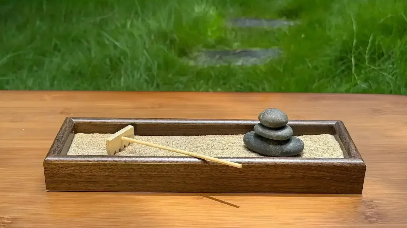 Wooden tabletop garden with three stacked river stones, sand, and a bamboo rake on an outdoor table.