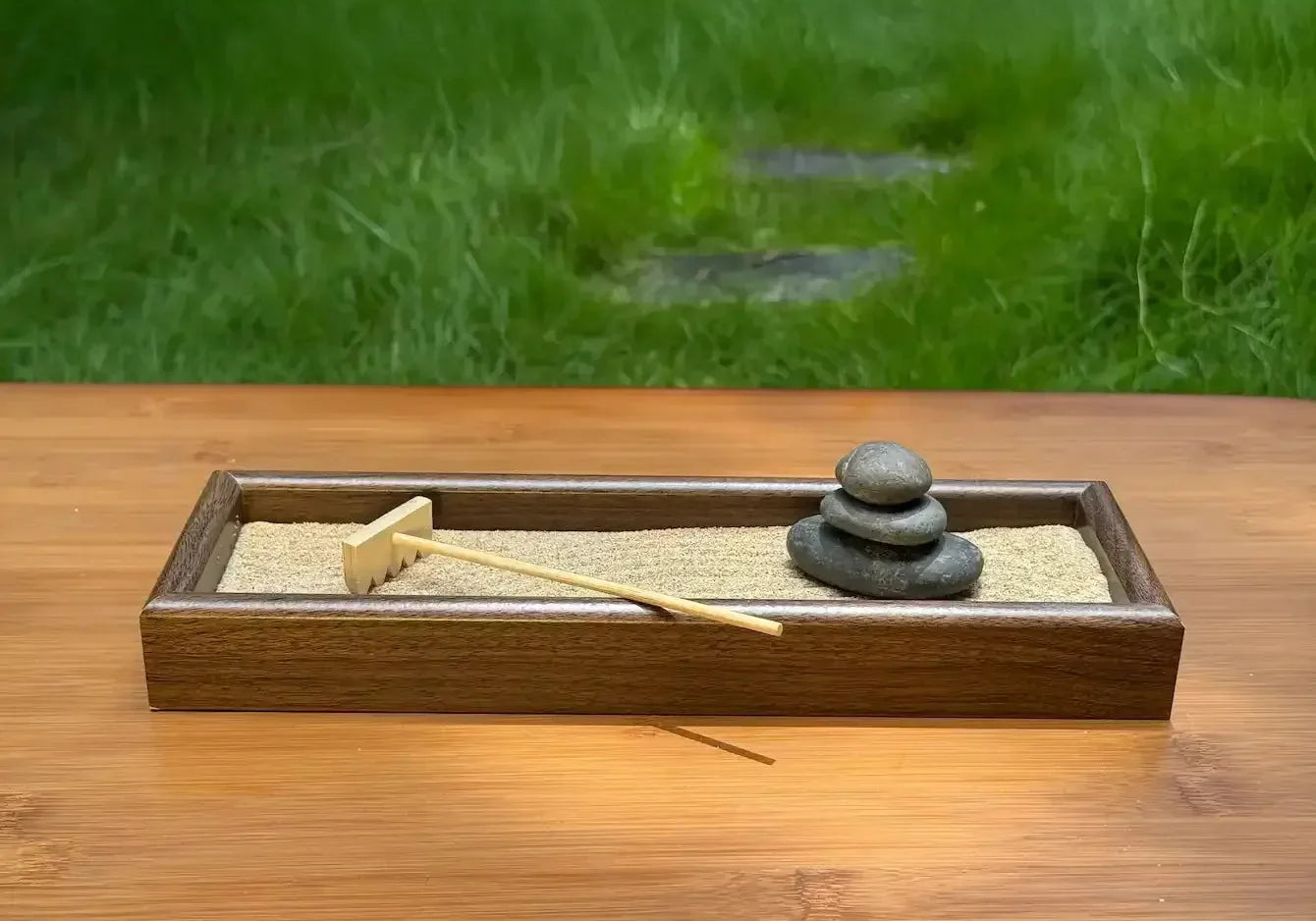 Wooden tabletop garden with three stacked river stones, sand, and a bamboo rake on an outdoor table.