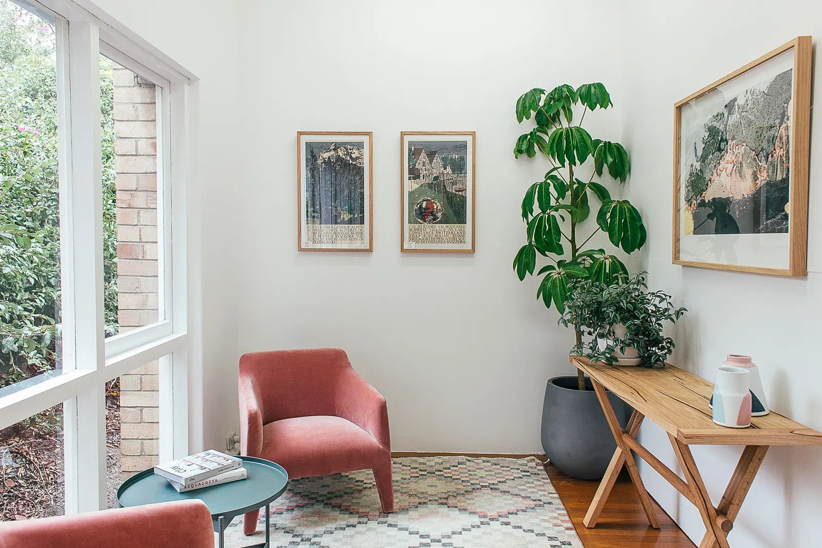 A pink armchair beside large windows, indoor plants, and a wooden console table.