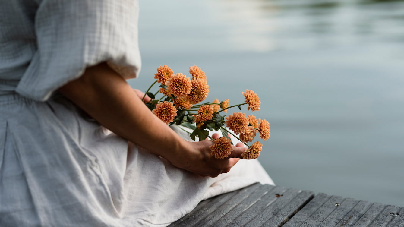A woman in a white dress holding a bouquet of orange flowers by a calm lake, reflecting the truth about energy.