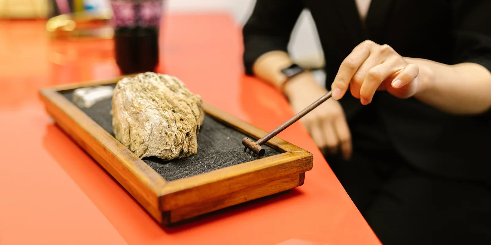 A hand guides a small wooden rake through dark sand around a stone in a tabletop garden on a red desk.