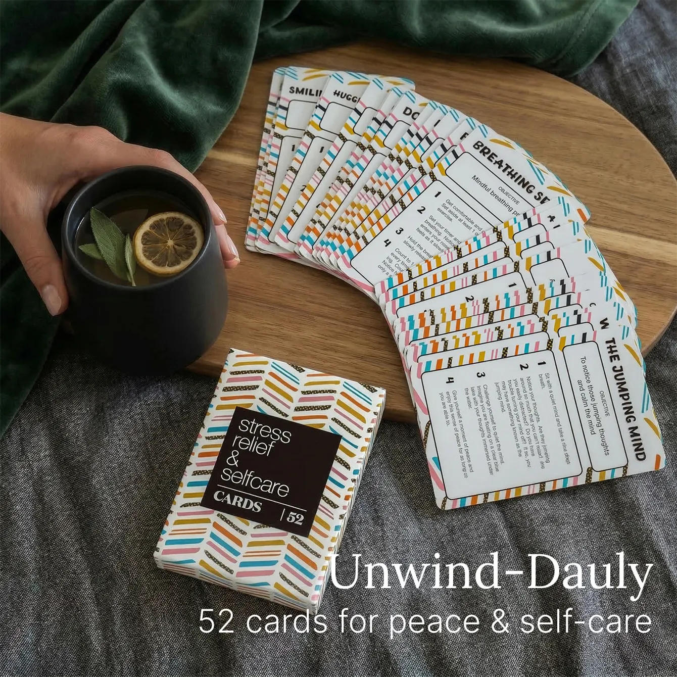 A person holding a mug with a lemon slice beside a fan of 52 Self Care Cards on a wooden surface.
