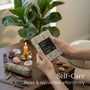 Woman holding Self Care Cards on a wooden table with candles and crystals, promoting mindfulness and relaxation.