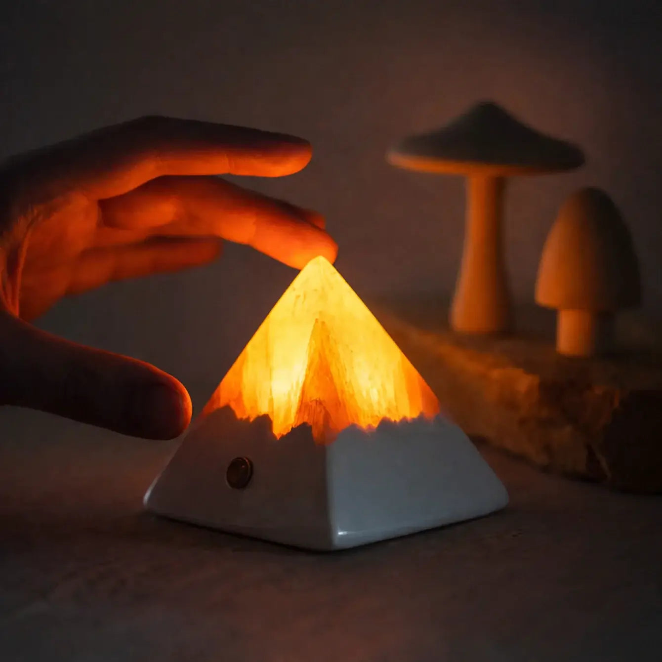 Hand adjusting the Mineral Pyramid Stone Lamp emitting a warm glow, enhancing the ambiance of the surrounding space.