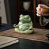 Large green glazed ceramic meditation frog beside a tea cup on a brown cloth.