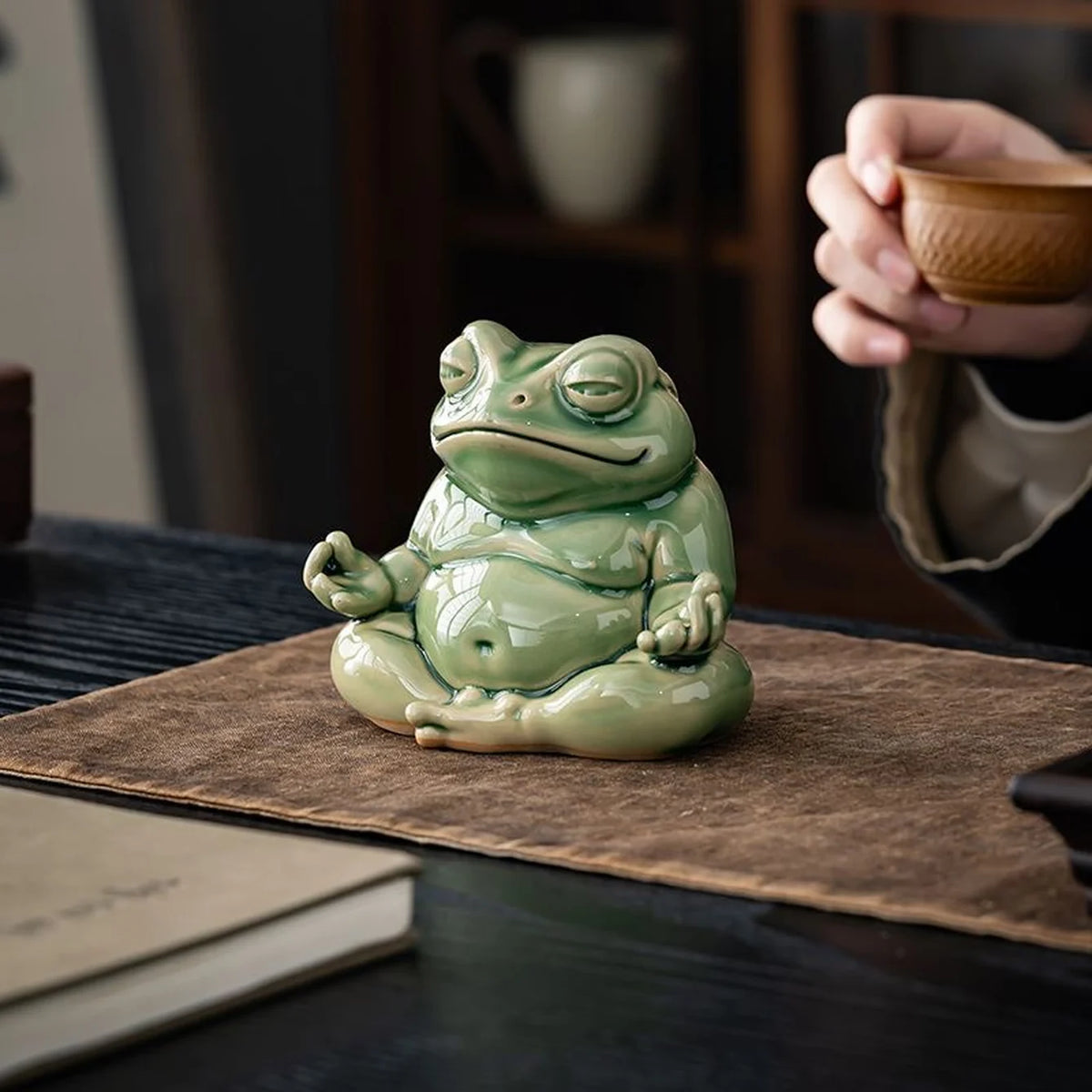 Large green glazed ceramic meditation frog beside a tea cup on a brown cloth.