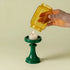 Hand pouring a yellow liquid from a jar into a green candle holder with a lit candle inside on a beige background.