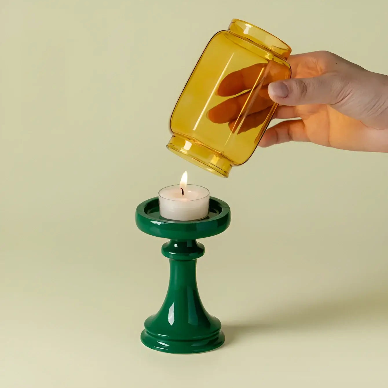 Hand pouring a yellow liquid from a jar into a green candle holder with a lit candle inside on a beige background.