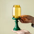 Hand holding a yellow glass container on a green stand against a beige background