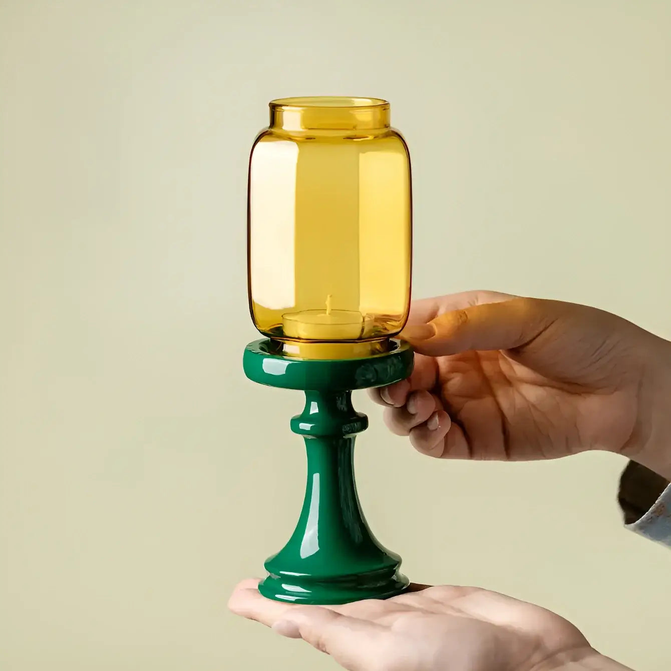 Hand holding a yellow glass container on a green stand against a beige background