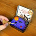 Hand arranging the miniature broom in a Halloween Crystal Zen Garden with purple sand, obsidian tree, raven, pumpkin, and stones.