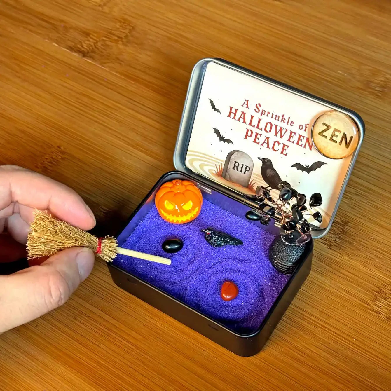 Hand arranging the miniature broom in a Halloween Crystal Zen Garden with purple sand, obsidian tree, raven, pumpkin, and stones.