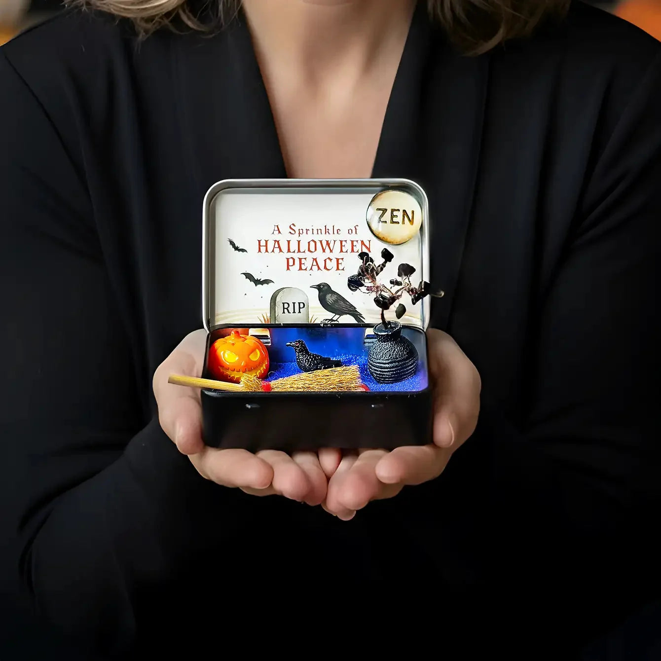 Person holding an open Halloween Crystal Zen Garden tin with purple sand, obsidian tree, raven, pumpkin, and miniature broom.