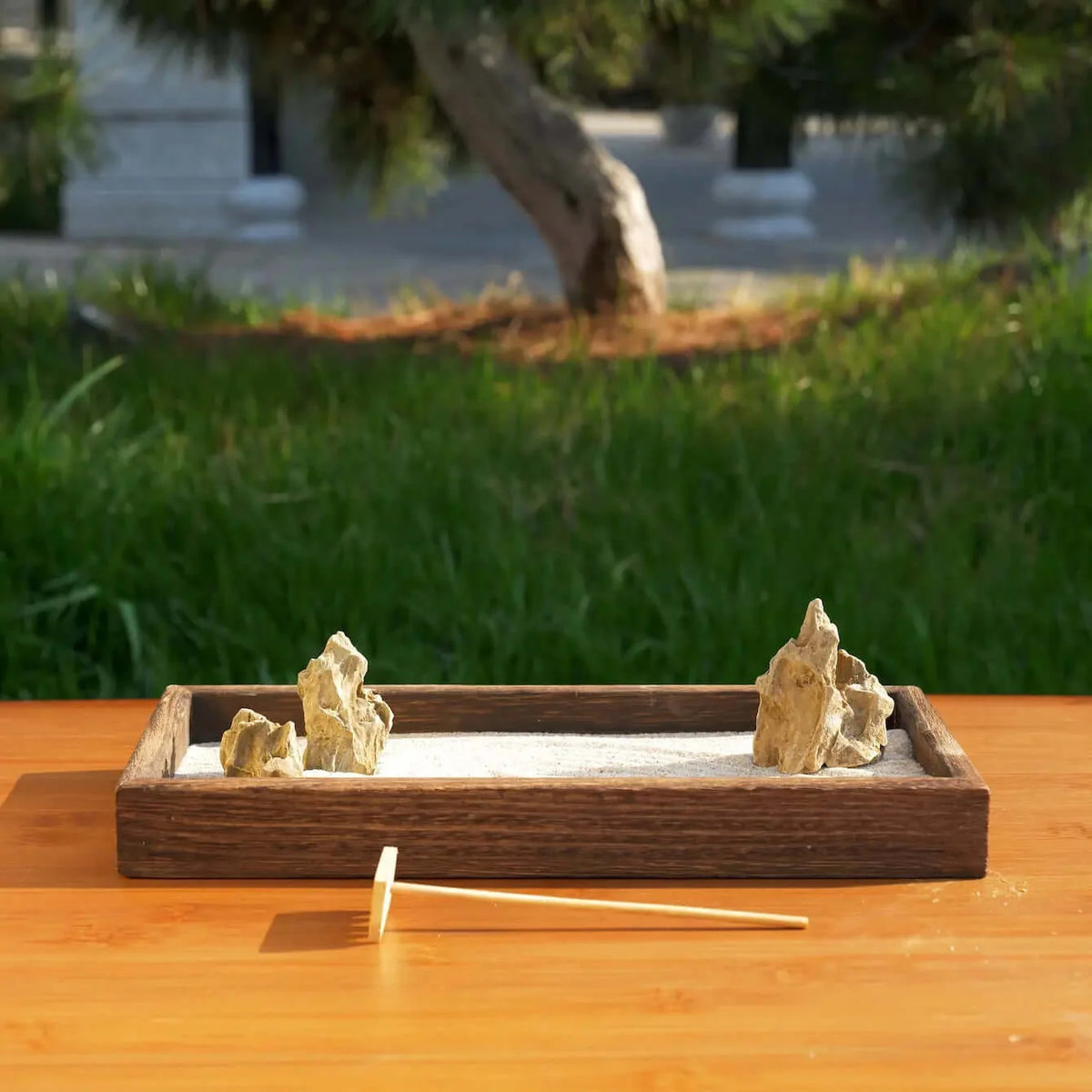 Japanese Karesansui Zen Garden outdoors with natural stones, raked white sand, dark wood tray, and wooden rake.