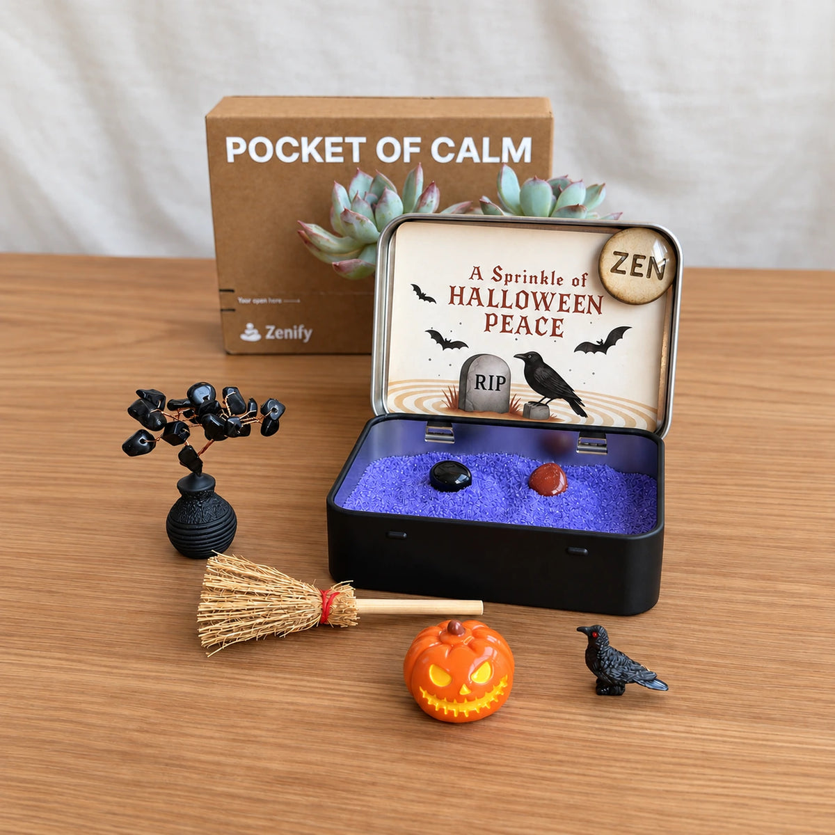 Classic Halloween Zen Garden on a wooden desk with purple sand, obsidian tree, pumpkin, raven and miniature broom arranged beside the tin.