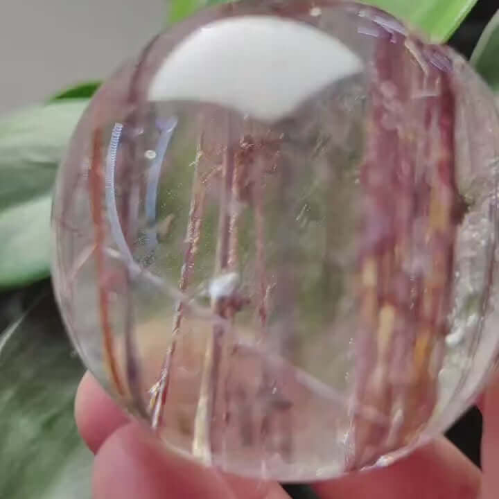 Zenify™ Rutilated Quartz Crystal Sphere, 42mm, showcasing golden rutile threads, held in hand with greenery in background.