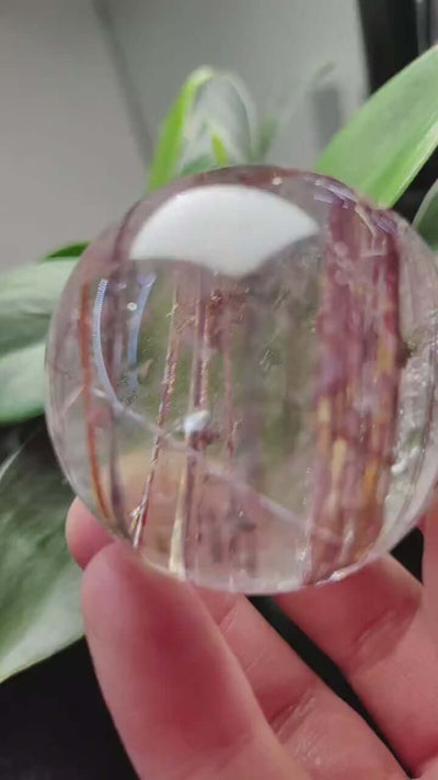 Zenify™ Rutilated Quartz Crystal Sphere, 42mm, showcasing golden rutile threads, held in hand with greenery in background.