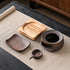 Wooden base, round ceramic warmer body, curved dish, and handled candle cup arranged separately on a woven mat.