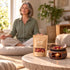 A softly blurred mature woman meditates beside a Soft Pause 20 g botanical blend pouch and a lit ceramic warmer holding rose petals and orange peel.