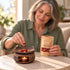 Shallow-focus scene of a mature woman with rose petals and orange peel beside a Soft Pause 20 g pouch and a ceramic warmer with a lit tealight.