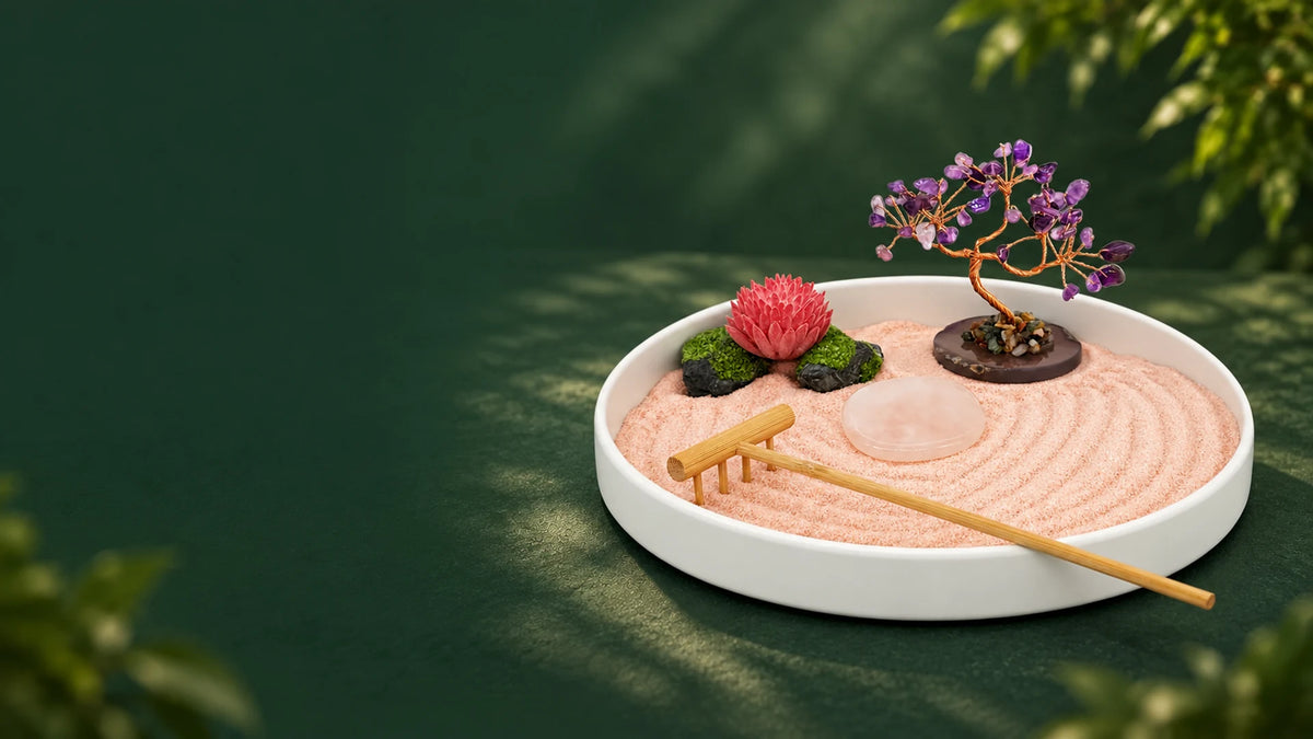 A white round Zen garden tray with peach-pink sand, a purple crystal tree, pink lotus ornament, pale pink polished stone, and bamboo rake on a forest-green surface; landscape banner.