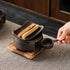 A hand displays the empty removable ceramic candle cup beside the warmer on its wooden base, with wood pieces on the dish.