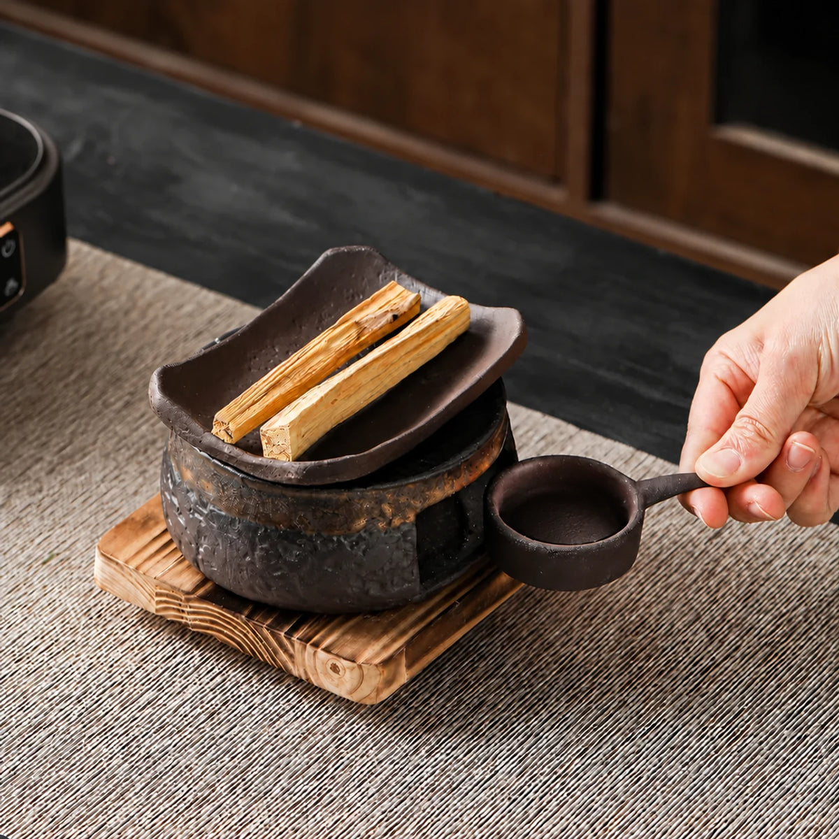 A hand displays the empty removable ceramic candle cup beside the warmer on its wooden base, with wood pieces on the dish.